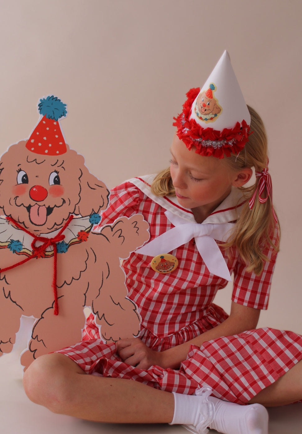 Child in a red checkered dress with a clown hat sitting next to a paper cutout of a clown dog.